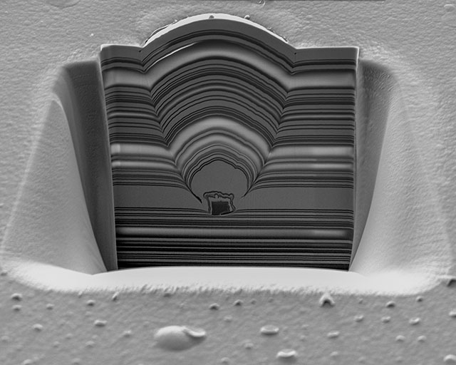 FIB-Querschnitt durch eingewachsenen Partikel. Optisches Multilagen-Schichtsystem aus sich abwechselnden SiO2- und Ta2O5-Schichten. Unter der Oberfläche ist ein überwachsener Si-Partikel von ca. 2 µm Durchmesser zu erkennen.
