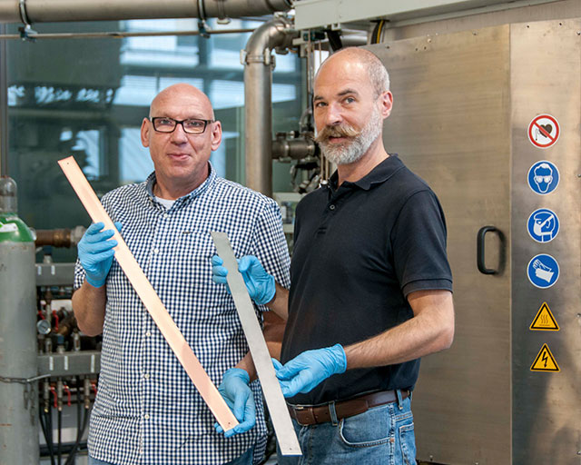 The development engineers of the IST in front of the coating plant: Ralf Wittorf (left) with a copper-coated pattern of titanium, beside him Torsten Hochsattel with the untreated sample material.